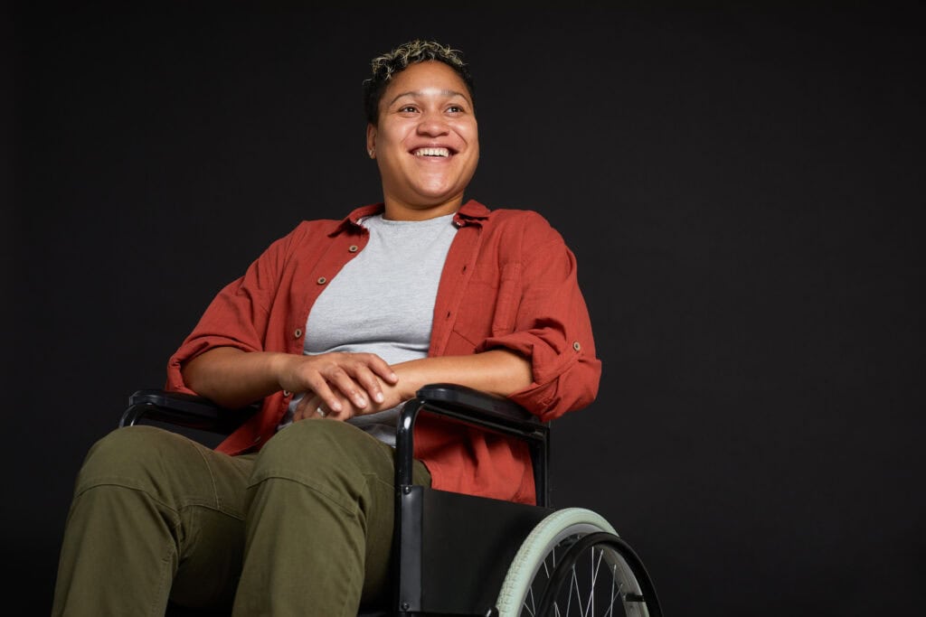 Young woman with disabilities sitting in wheelchair and smiling isolated on black background