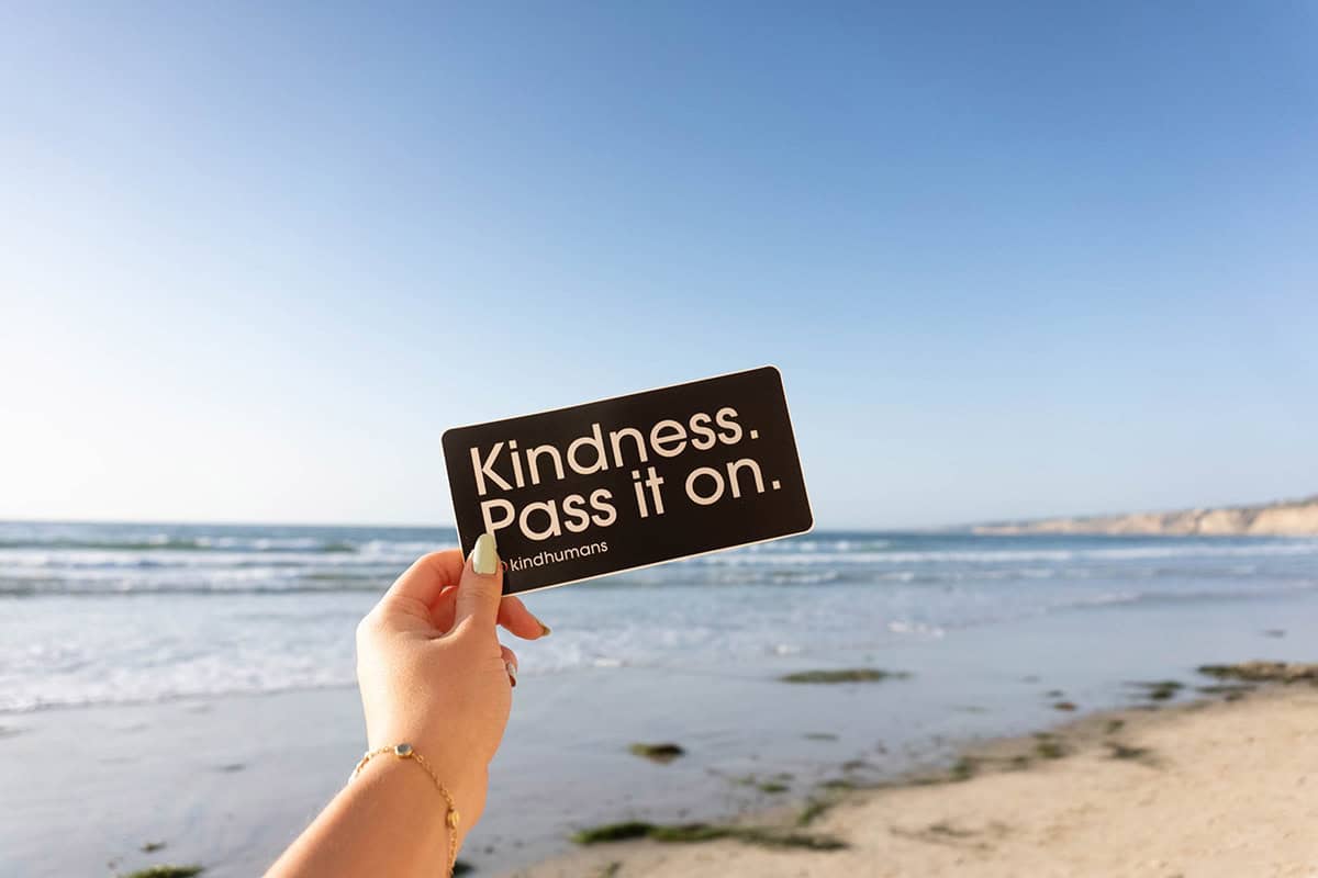 Hand holding a card with 'Kindness. Pass it on.' at the beach.